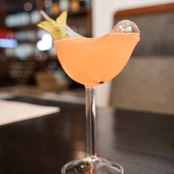 A vibrant orange cocktail in a coupe glass, garnished with mint and a clear ice sphere.