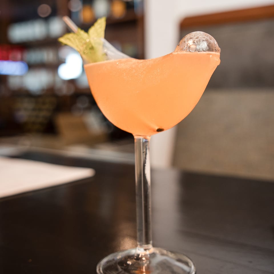 A vibrant orange cocktail in a coupe glass, garnished with mint and a clear ice sphere.
