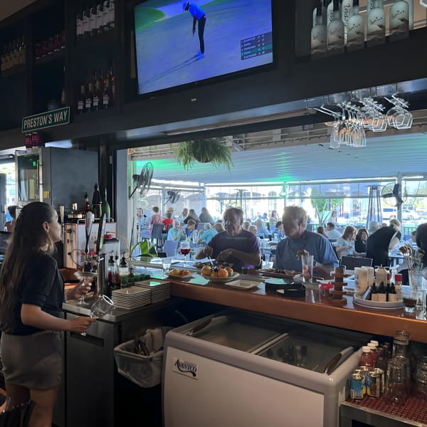 A bartender prepares drinks at a lively bar with patrons dining and watching golf on TV.